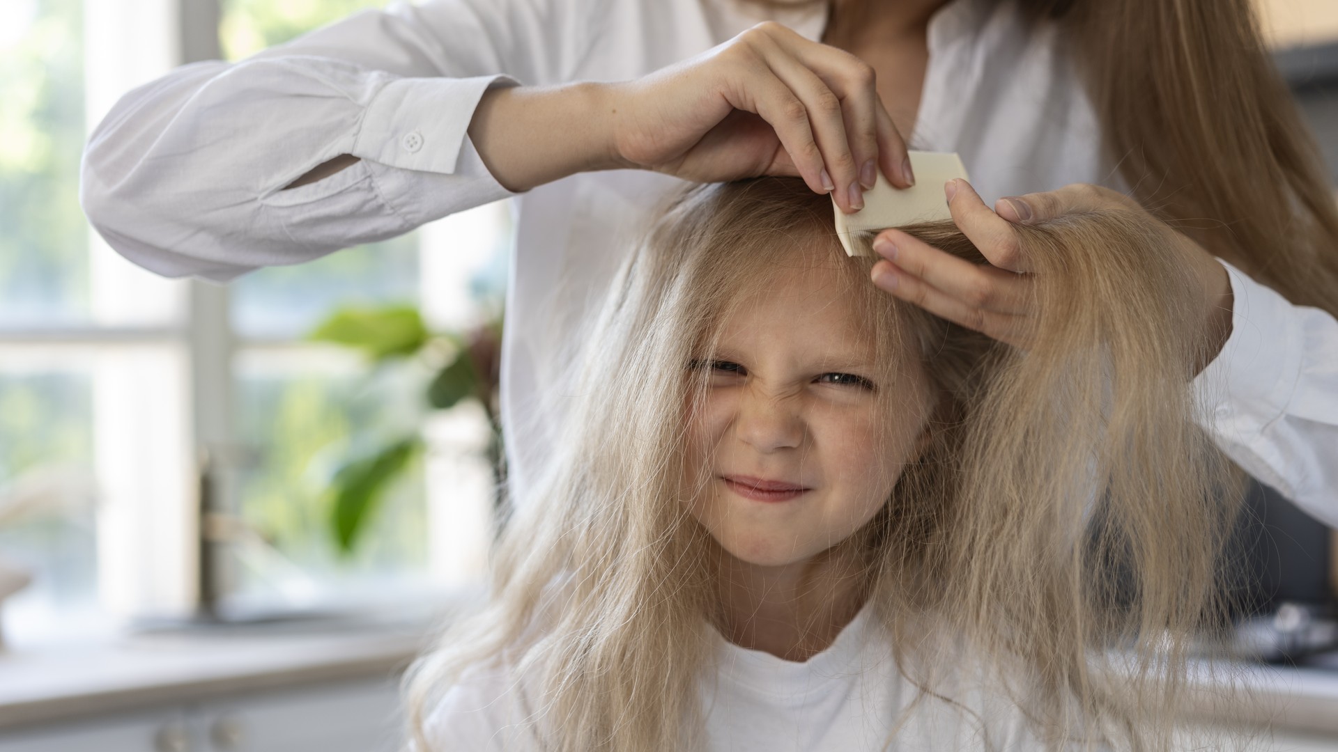 A parent parts a section of a young child's fine blonde hair near the nape to check the scalp under bright natural light.