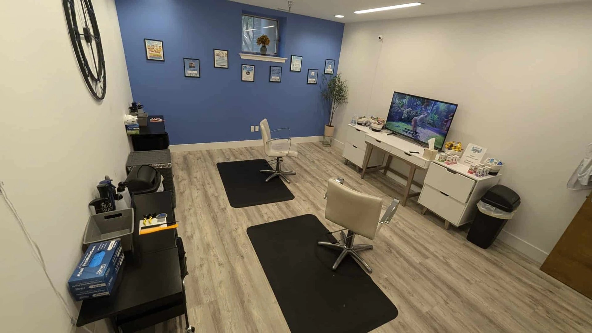 Treatment room with two white salon chairs on black floor mats, a wall-mounted television and a white console table with supplies beside a blue accent wall